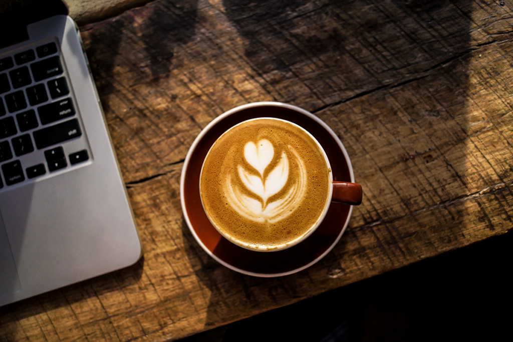 Laptop agus cupán caife ar dháileog adhmaid i ngníomhaireacht dearaidh gréasáin i mBAC.