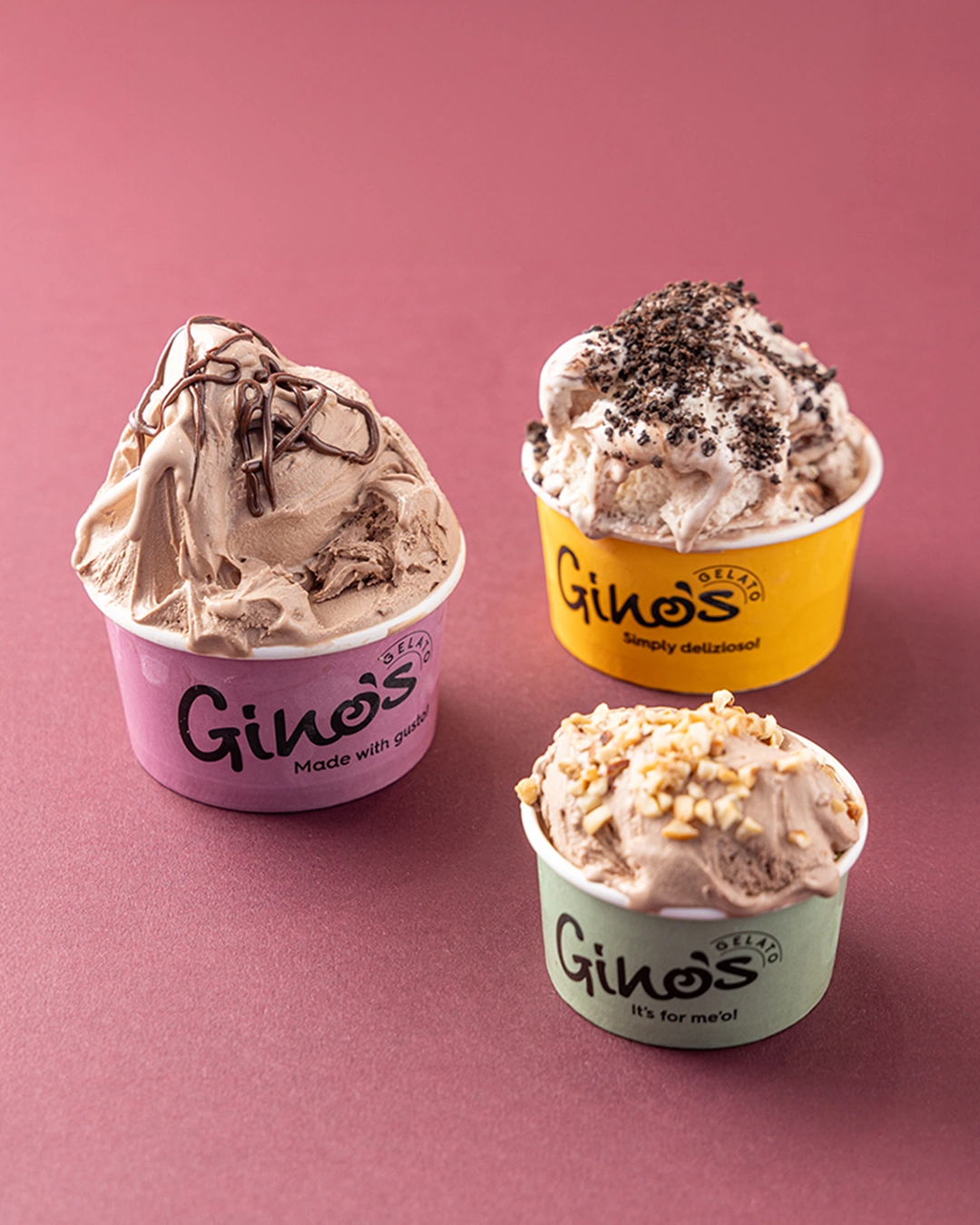 Three cups of Gino's gelato on a pink background. Various flavours topped with chocolate and nuts. Delicious treat.
